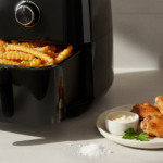 Schwarzer Airfryer mit Pommes, daneben Hähnchenflügel und Dip.
