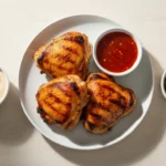 Gegrillte Hähnchenkeulen mit Dip-Saucen auf weißem Teller.