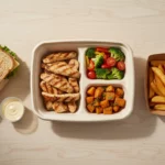 Mittagessen-Box mit gegrilltem Hähnchen, Gemüse und Pommes.