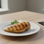 Gegrilltes Hähnchenbrustfilet mit Zitronenscheibe und Petersilie auf Teller, Smartphone daneben.