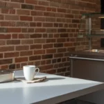 Weiße Tischplatte mit Metallbox und Kaffeebecher vor Backsteinwand.